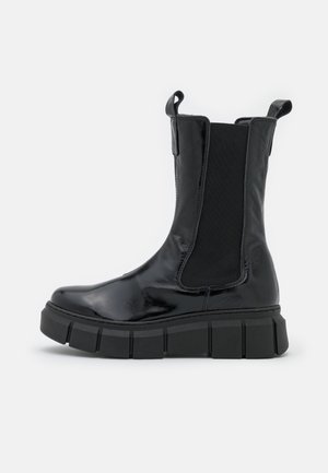 Botte d'ankle en cuir noir avec semelle épaisse, panneaux latéraux élastiques et languettes au sommet sur fond blanc.