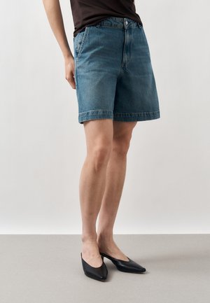 Personne portant un short en denim bleu mi-long, des talons noirs à bout pointu et un haut noir, debout devant un fond clair uni.