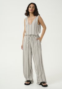Combinaison rayée en beige clair et gris foncé. Elle a un décolleté en V profond, un design sans manches, une taille élastique et deux poches avant.