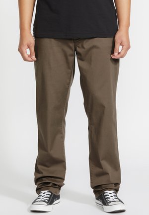 Personne portant un pantalon droit marron, des baskets basses noires avec des lacets blancs et une chemise noire, debout les mains détendues le long du corps.