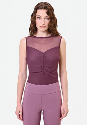 Donna con capelli castani mossi che indossa una canotta viola senza maniche con pannello superiore trasparente e pantaloni a vita alta color malva, in piedi davanti a uno sfondo semplice.