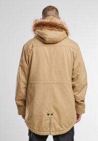 Brun parka-jakke med hette foret med pels, synlig søm, bakventiler og trykknapper ved kanten. Enkelt design og funksjonelt utseende.