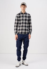 Camisa a cuadros en blanco y negro, pantalones cargo azul marino y gorra de béisbol gris. Las zapatillas tienen detalles en blanco y gris. Atuendo casual en modelo.