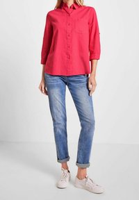Chemise rouge à boutons avec une poche poitrine et des manches trois-quarts, associée à un jean bleu clair retroussé et des baskets blanches.