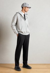 Um hoodie cinza com fecho éclair sobre uma camisa branca com riscas e uma gravata preta, combinado com calças pretas e sapatilhas pretas, contra um fundo neutro.