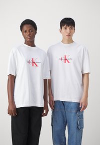 Bílé bavlněné trička s logem "Calvin Klein Jeans" v červené barvě, krátké rukávy, volný střih. Spárované s černými kalhotami a modrými kapsáči.