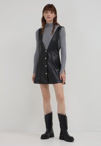 Frau, die einen grauen Rollkragenpullover trägt, kombiniert mit einem schwarzen ärmellosen Lederkleid und schwarzen kniehohen Stiefeln, steht vor einem schlichten Hintergrund.