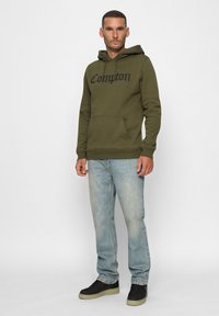 Hombre con sudadera oliva "Compton", jeans azul claro y zapatillas negras, de pie contra un fondo blanco.
