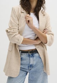 Jasnobeżowa lniana marynarka z podwiniętymi rękawami, z kieszeniami i luźnym krojem, noszona na białym prążkowanym topie i niebieskich dżinsach z czarnym paskiem.