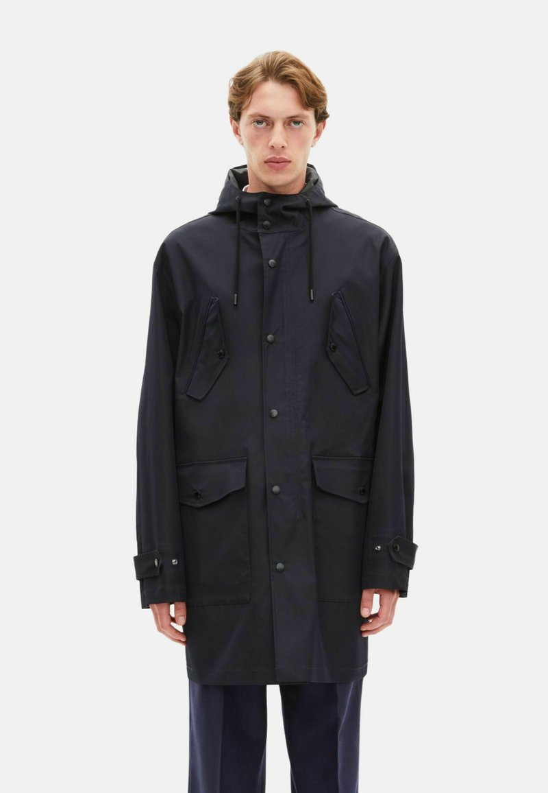 Parka imperméable navy avec capuche, quatre poches, boutons-pression et cordons de serrage. Présente une texture lisse et des poignets ajustables.