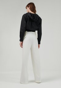 Chaqueta negra corta con capucha que cae sobre pantalones anchos blancos con bolsillos rectangulares y una textura suave.