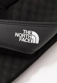 Czarny klapek z teksturowanym wierzchem, na którym znajduje się biały napis "The North Face", oraz gładką, matową czarną podeszwą.