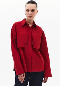 Camicia rossa a maniche lunghe con colletto a camicia, due grandi tasche anteriori e maniche ampie. Realizzata in tessuto morbido con texture minima.