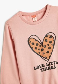 Roze sweatshirt met ronde hals, voorzien van een grote bruine hartafbeelding met zwart bloemenpatroon en de tekst "HOUD VAN KLEINE DINGEN" eronder.