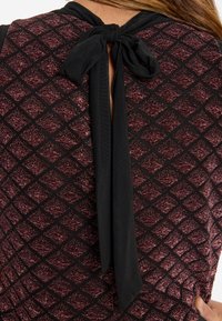 Zwarte top met een gestructureerd bordeauxrood ruitpatroon, voorzien van een zwarte strik aan de achterkant en een sleutelgatopening eronder.