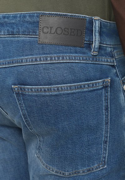 Denim jeans in medium blue with a dark leather logo patch labeled "CLOSED." Features two back pockets with contrasting stitching details.
