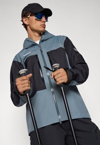 Blue and black outdoor jacket with a zippered front, two chest pockets, and long sleeves. Paired with trekking poles and sunglasses.