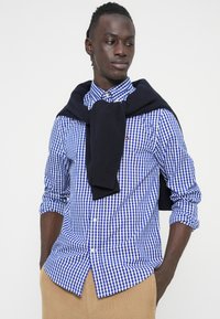 Ανδρικό πουκάμισο με μπλε και λευκό καρό, κουμπωτό με μακριά μανίκια, συνοδευόμενο από μπλε σκούρα μπλούζα δεμένη στους ώμους και μπεζ παντελόνι.