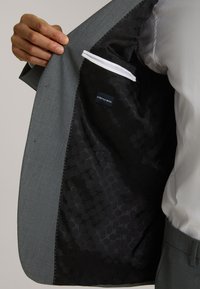 Grau dehnbarer Wollanzugjacke Innenleben mit einem schwarzen gemusterten Futter. Verfügt über eine einzelne weiße Tasche und subtile weiße Nähte.