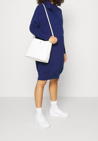 Borsa bianca con bordo nero, caratterizzata da un design semplice e una forma strutturata, abbinata a un vestito a maglia blu navale e sneakers bianche.