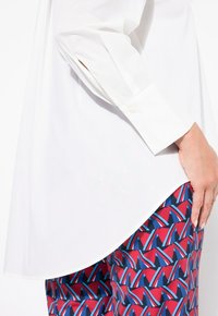 Chemise blanche à boutons avec des manches longues, assortie à un pantalon à motifs comportant des formes géométriques rouges, bleues et noires. Texture de tissu lisse.