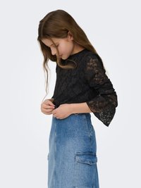 Juoda nėriniais puošta ilgomis rankovėmis palaidinė su varpelio formos rankovėmis, derinama su šviesiai mėlynos spalvos džinsine sijonu su šoninėmis kišenėmis ir aukštu liemeniu.