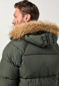 Homme dans une veste en duvet verte avec une capuche doublée de fourrure, de profil, montrant son dos et son profil latéral sur fond uni.