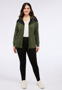 Felpa con zip di colore verde oliva con dettagli in pizzo nero, maniche lunghe e cappuccio con cordino, abbinata a pantaloni neri e sneakers bianche.