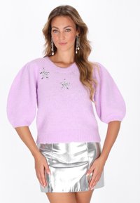 Lila gestrickter Pullover mit kurzen Puffärmeln, versehen mit zwei dekorativen Stern-Applikationen. Kombiniert mit einem metallisch silbernen Minirock.