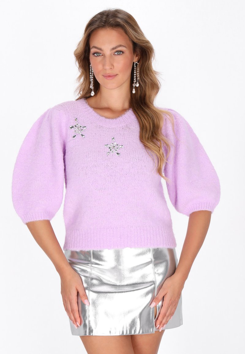 Lila gestrickter Pullover mit kurzen Puffärmeln, versehen mit zwei dekorativen Stern-Applikationen. Kombiniert mit einem metallisch silbernen Minirock.