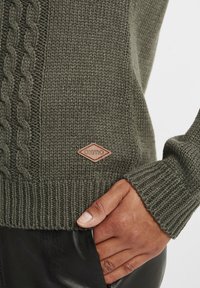 Dark green knitted sweater with cable pattern, ribbed cuffs, and hem. Features a branded leather patch on the front. Soft texture.