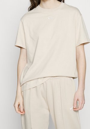 Personne portant un t-shirt Nike beige et un pantalon assorti avec de petits logos Nike blancs sur la poitrine et la cuisse gauche, debout devant un fond uni.