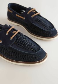 Chaussures bateau en cuir tressé bleu marine avec lacets marron clair et semelles en caoutchouc blanches sur un fond neutre.