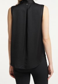 Blouse noire en satin sans manches avec un pli au dos et un ourlet arrondi portée par une personne en pantalon noir, vue de dos.