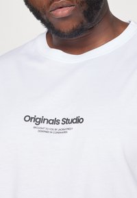 Balta medvilninė marškinėliai su apvaliu iškirpimu ir juodu spaudiniu: "Originals Studio Brought to You by JACK & JONES Designed in Copenhagen."