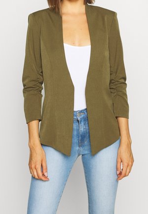 Vrouw met een olijfgroene open blazer met geraffelde mouwen, wit topje en lichtblauwe spijkerbroek, staand tegen een effen achtergrond.