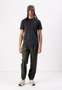 Schwarzes T-Shirt, lockere Passform; olivgrüne Hose, mit elastischen Bündchen; beigefarbene Sneakers; gemusterte Kappe, flacher Schirm.
