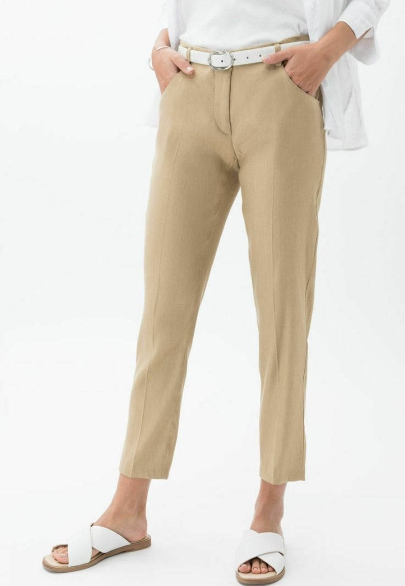 Pantaloni beige su misura con vestibilità slim, caviglie orlate, tasche laterali e una cintura bianca con una fibbia circolare argentata. Indossati con sandali bianchi.