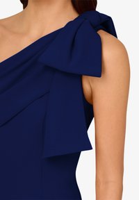 Námořnická modrá šaty s drapovaným výstřihem a velkou mašlí na levém rameni. Hladká látka, přiléhavý střih a bezrukávový design.