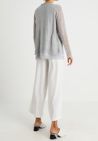 Jasnoszary koronkowy kardigan z długimi rękawami i wzorzystym dołem, założony na szary top, zestawiony z białymi szerokimi spodniami i białymi sukiennymi klapkami.