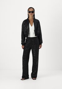 Black sequined bomber jacket with fitted cuffs, paired with matching black sequined wide-leg pants and a white v-neck top. Black sunglasses worn.