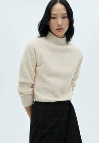 Maglione beige chiaro e morbido con collo alto, maniche raglan e polsini a coste; abbinato a una gonna a quadri scura con dettagli verdi.