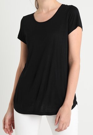 Kvinde iført en helt sort t-shirt med korte ærmer og hvide bukser mod en lys baggrund.