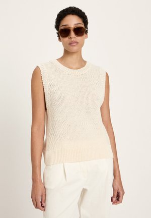 Person trägt ärmelloses cremefarbenes Strickoberteil, weiße Hose und große braune Sonnenbrille, steht vor einem schlichten hellen Hintergrund.