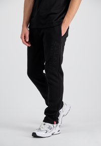 Sorte sweatpants lavet af blødt materiale, med et fremtrædende præget "ALPHA INDUSTRIES" logo på siden. Stylet med hvide sneakers.