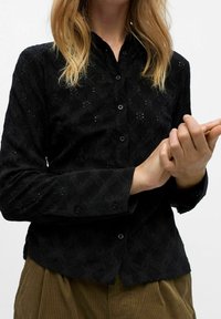 Chemise noire à boutons avec un motif en losanges texturé, présentant un col arrondi et des boutons sur le devant, portée avec un pantalon beige.