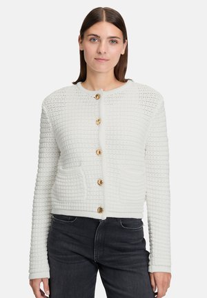 Femme portant un cardigan en tricot texturé blanc avec des boutons dorés et des poches avant, associé à un jean noir, se tenant devant un fond uni.