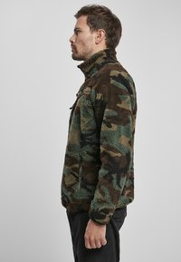 Chaqueta de forro polar de camuflaje con alto cuello, con patrones en verde, marrón y negro. Incluye bolsillos con cremallera lateral y tejido texturizado.