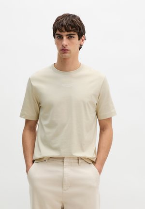 Junger Mann trägt beige T-Shirt mit kurzen Ärmeln und passende Hose, steht mit den Händen in den Taschen vor einem schlichten weißen Hintergrund.