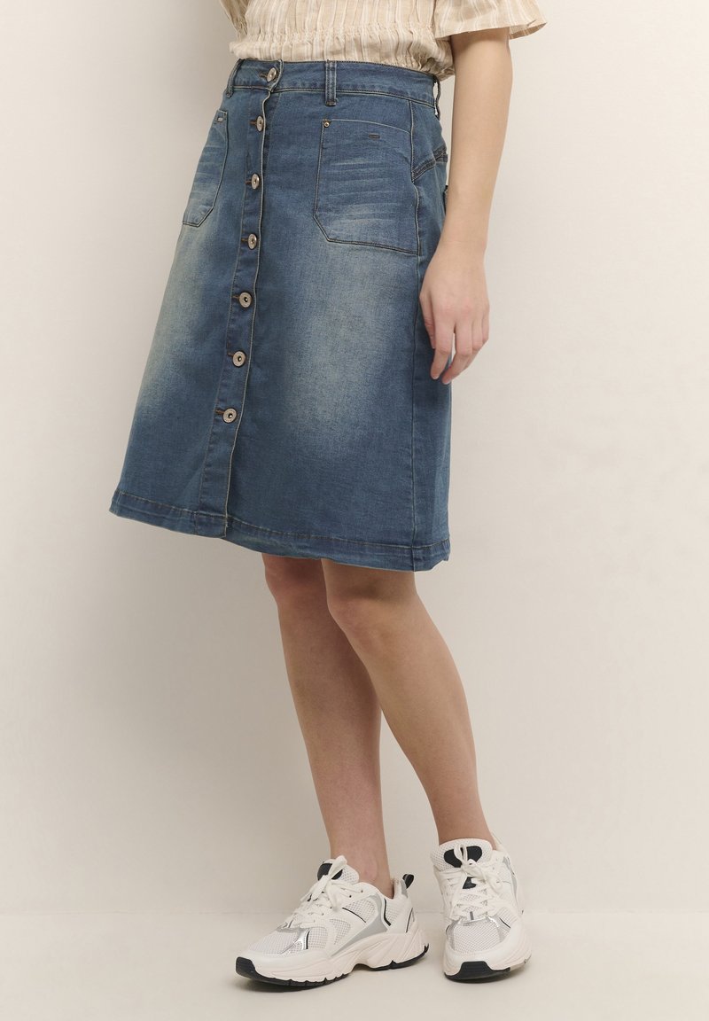 Kvinna klädd i en knälång blå denimkjol med framknappar och fickor, tillsammans med vita sportskor, stående mot en neutral bakgrund.